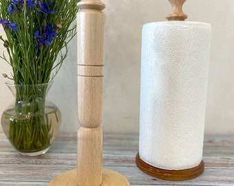 May include: A wooden paper towel holder with a natural finish. The holder has a round base and a tall, cylindrical post with a knob on top. A roll of white paper towels is on the holder.