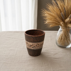 Può includere: Un bicchiere in ceramica marrone fatto a mano con una fascia decorativa con un motivo floreale color crema. Il bicchiere ha un aspetto rustico e viene probabilmente utilizzato per bere bevande. L'interno del bicchiere è di colore marrone scuro.