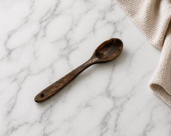 May include: A dark brown wooden spoon with a rounded bowl and a long handle. The spoon has a small hole at the end of the handle. A beige cloth is in the background. The spoon is on a white marble surface.