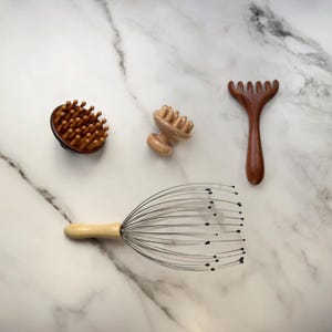May include: A collection of four massage tools including a round wooden scalp massager with multiple prongs, a small wooden knob massager, a wooden rake-style massager, and a metal wire head massager with a light-coloured wooden handle.
