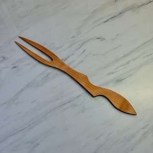 Op de afbeelding: Een houten vleesvork met twee tanden en een gebogen handvat. De vork is gemaakt van lichtgekleurd hout en heeft een gladde afwerking. De handgreep heeft een decoratieve vorm. De vork is ontworpen om te serveren.