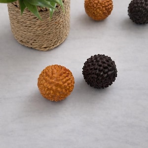 Pode incluir: Close-up de esferas decorativas com superfícies texturizadas. Uma esfera é laranja e a outra marrom escuro. As esferas têm um padrão irregular e irregular. Uma cesta tecida com uma planta verde está no fundo.