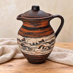 May include: A round clay pitcher with a matching lid, featuring a dark brown and terracotta colour palette. The body is decorated with horizontal bands and a central section displaying a black leaf pattern on a cream-coloured background.