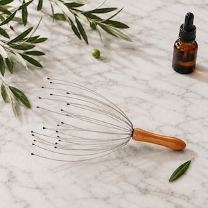 May include: A scalp massager featuring a smooth, light brown wooden handle and multiple thin, flexible metal wires that fan out, each tipped with a small black bead. Beside it sits a small, dark amber glass dropper bottle with a black cap.