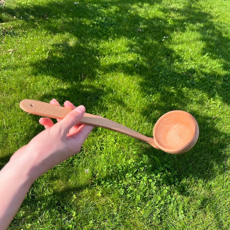 May include: A wooden ladle with a long handle and a round bowl. The ladle is made of light-colored wood and has a hole in the handle for hanging. The background is a green grassy area.