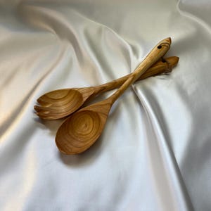 Könnte beinhalten: Zwei Salatbestecke aus Holz, ein Löffel und eine Gabel, werden auf einer weißen Satin-Oberfläche präsentiert. Der Löffel hat eine abgerundete Schüssel, während die Gabel drei Zinken hat. Die Holzmaserung ist sichtbar, und die Farbe ist hellbraun.