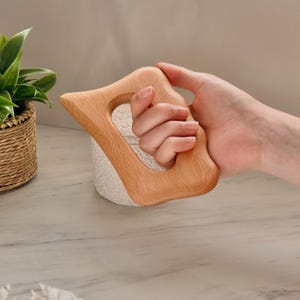Puede incluir: Una herramienta de masaje de madera clara con un mango curvo y una abertura central. La herramienta se sostiene en una mano, mostrando su diseño ergonómico. La veta de la madera es visible, destacando el material natural.
