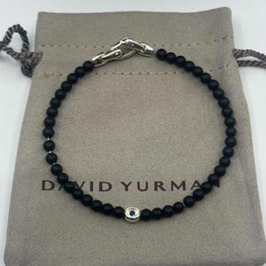 May include: Black beaded bracelet with a silver clasp and a small silver charm with a blue center. The bracelet is displayed on a gray pouch with the brand name "DAVID YURMAN" printed on it.