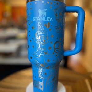 Pode incluir: Uma caneca de viagem de aço inoxidável azul com alça. A caneca apresenta um design prateado de Mickey Mouse como mágico, estrelas e o logotipo da Stanley.