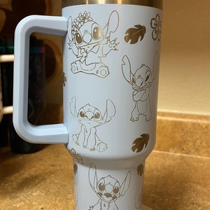 Op de afbeelding: Een lichtblauwe reistumbler van roestvrij staal met een zwarte handgreep en een deksel. De tumbler heeft een herhalend patroon van Stitch uit Lilo & Stitch in verschillende poses.