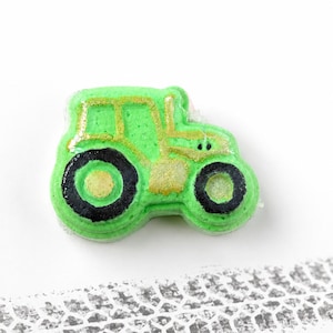 Puede incluir: Una bomba de baño con forma de tractor de color verde brillante, con neumáticos negros de centros dorados y detalles dorados en la carrocería. Debajo del tractor, un patrón de huella de neumático gris imita las marcas de un vehículo.