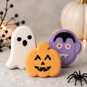 May include: Three bath bombs shaped like Halloween characters: a white ghost with black eyes and mouth, an orange pumpkin with a black jack-o'-lantern face, and a purple vampire with black hair, red eyes, and white fangs, arranged together.