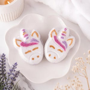 Puede incluir: Dos bombas de baño blancas con forma de unicornio, con orejas, ojos y fosas nasales pintados en dorado, y un detalle de crin rosa y morado en la frente. Las bombas de baño están colocadas una al lado de la otra en un plato de cerámica blanco.