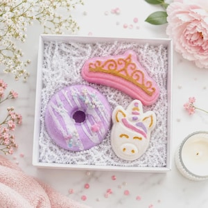 Puede incluir: Una caja de regalo blanca que contiene tres bombas de baño: una tiara rosa con detalles dorados, una dona morada con glaseado blanco y chispas, y una cabeza de unicornio blanca con crin rosa y morada, sobre una base de papel triturado.