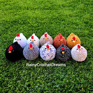 May include: A collection of ten plush, round chicken figures with red combs and yellow beaks, each in a different color, including white, black, gray, and brown. The chickens are arranged on a bed of green grass.