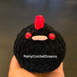 May include: A round, black crocheted plush toy shaped like a chicken, featuring a bright red crest on top, two shiny red plastic eyes, and a small white beak. The text RainyCrochetDreams is printed in white across the center of the black body.