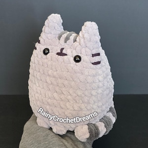 May include: A light gray crocheted cat plush toy with a soft, textured yarn finish. The cat features dark gray stripes on its head and tail, black plastic eyes, and a small embroidered mouth. The text RainyCrochetDreams is visible on the front.