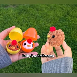 May include: A close-up of three crochet chickens, two inside orange and yellow plastic eggs, and one held separately. The chickens are brown with red combs and black eyes. The text "RainyCrochetDreams" is visible.