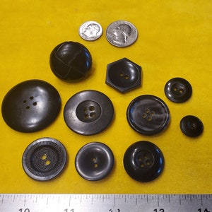 May include: A collection of 11 black buttons of various shapes and sizes, including round, hexagonal, and a button with a textured surface. The buttons are arranged on a yellow surface with a ruler in the bottom of the image.