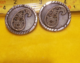 May include: Two round, silver-toned earrings with a paisley design. The center of each earring features a gold-colored paisley with dark brown accents. The earrings are set against a yellow background.