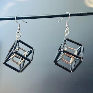 以下が含まれることがあります： シルバーのフックが付いた幾何学的なキューブイヤリング。イヤリングは、シルバーのインナーキューブデザインの黒いフレームが特徴です。グレーの背景に対して黒いロッドから吊り下げられています。