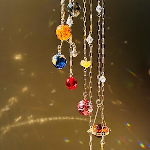 May include: A celestial-themed mobile featuring colorful, spherical beads representing planets, suspended from a gold-colored ring and chains. The planets are various colors, including blue, orange, red, and green, with clear crystal accents. A decorative piece for home decor.