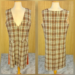 Op de afbeelding: Een vintage mouwloze jurk met een ruitpatroon in bruin-, crème- en zwarttinten. De jurk heeft een diepe V-hals, twee voorzakken en oranje zijpanelen. De jurk wordt op een mannequin gepresenteerd.