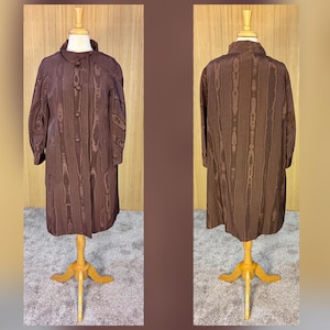 May include: A brown, knee-length coat with a textured, vertical stripe pattern. The coat has a button-up front and long sleeves. The coat is displayed on a wooden mannequin. The background is a light brown color.