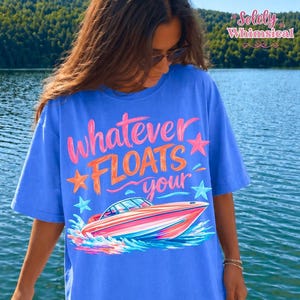 May include: A blue t-shirt with the phrase "Whatever Floats Your" and a graphic of a speedboat. The text is in pink and orange with star accents. The boat is pink, orange, and blue, with blue water around it.