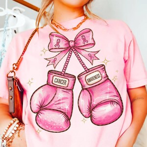 May include: Pink t-shirt featuring a graphic of pink boxing gloves with the words "Cancer" and "Awareness". The gloves are connected by a pink ribbon with pink ribbon accents. Gold glitter accents surround the gloves and ribbon.