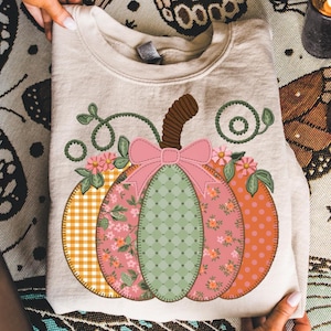 May include: Beige sweatshirt featuring a patchwork pumpkin design. The pumpkin has sections of orange polka dots, floral patterns, and green dotted fabric. A pink bow and brown stem complete the design. Perfect for fall.