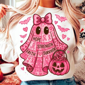 以下が含まれることがあります： ピンクのゴーストのデザイン、リボン、ハート、コウモリが特徴の白いスウェットシャツ。「希望、強さ、信仰、サバイバー」という言葉とピンクのリボンが描かれています。ピンクのジャック・オー・ランタンも特徴です。