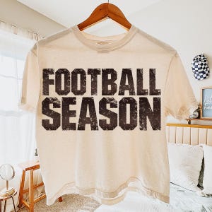 May include: A cream-coloured t-shirt with the words "FOOTBALL SEASON" printed in distressed black lettering. The shirt is hanging on a wooden hanger.