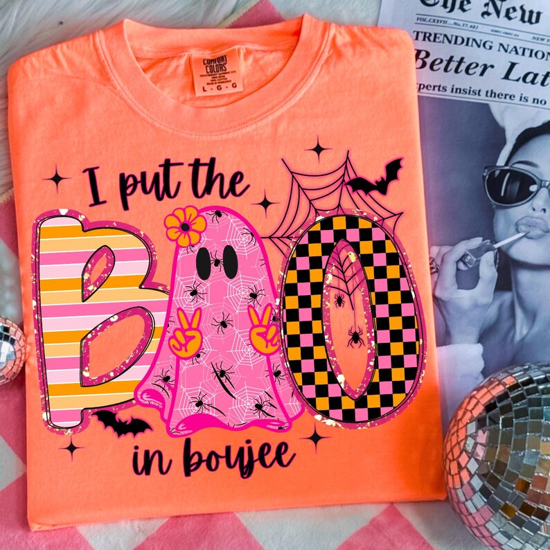 May include: An orange Comfort Colors t-shirt with the phrase "I put the BOO in boujee." The design features a pink ghost with peace signs, spiders, and a flower, along with a striped and checkered "BOO" design. A Halloween-themed graphic tee.