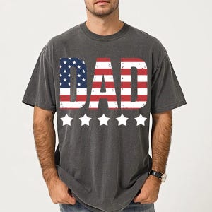 Puede incluir: Camiseta gris oscuro con la palabra "DAD" en rayas rojas y blancas y estrellas azules, que recuerda a la bandera americana. Cinco estrellas blancas est&aacute;n debajo. La camiseta es de estilo casual.