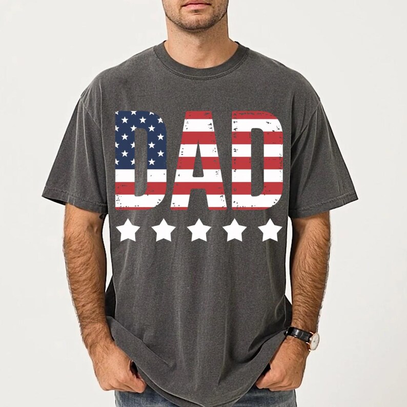 Puede incluir: Camiseta gris oscuro con la palabra "DAD" en rayas rojas y blancas y estrellas azules, que recuerda a la bandera americana. Cinco estrellas blancas est&aacute;n debajo. La camiseta es de estilo casual.