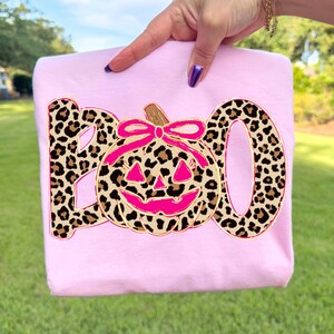 Op de afbeelding: Een lichtroze sweatshirt met het woord "BOO" in een luipaardprint. De "O" is vervangen door een roze pompoen met een bruine steel en roze strik. Het sweatshirt wordt door een hand omhoog gehouden.