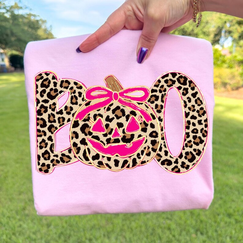 Op de afbeelding: Een lichtroze sweatshirt met het woord "BOO" in een luipaardprint. De "O" is vervangen door een roze pompoen met een bruine steel en roze strik. Het sweatshirt wordt door een hand omhoog gehouden.