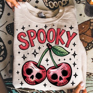 Może przedstawiać: Złożona beżowa bluza z napisem "SPOOKY" w czerwonych literach nad parą wiśni z czaszkami. Projekt zawiera zielone liście, łodygi i czarne akcenty gwiazd, tworząc motyw Halloween.