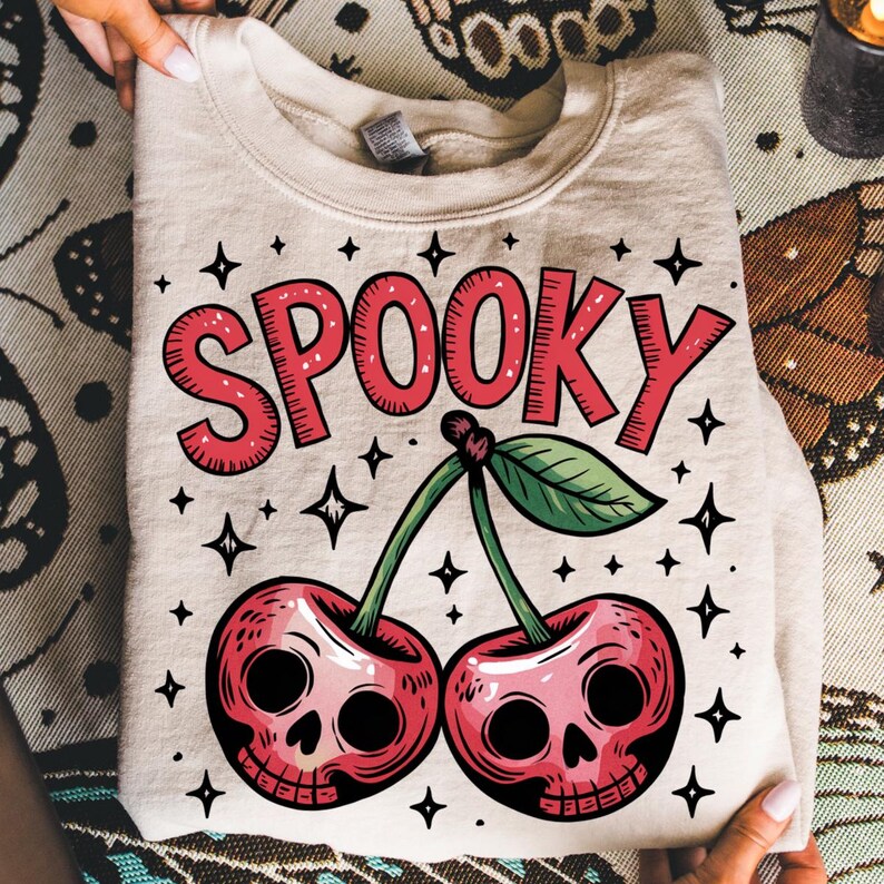 Może przedstawiać: Złożona beżowa bluza z napisem "SPOOKY" w czerwonych literach nad parą wiśni z czaszkami. Projekt zawiera zielone liście, łodygi i czarne akcenty gwiazd, tworząc motyw Halloween.