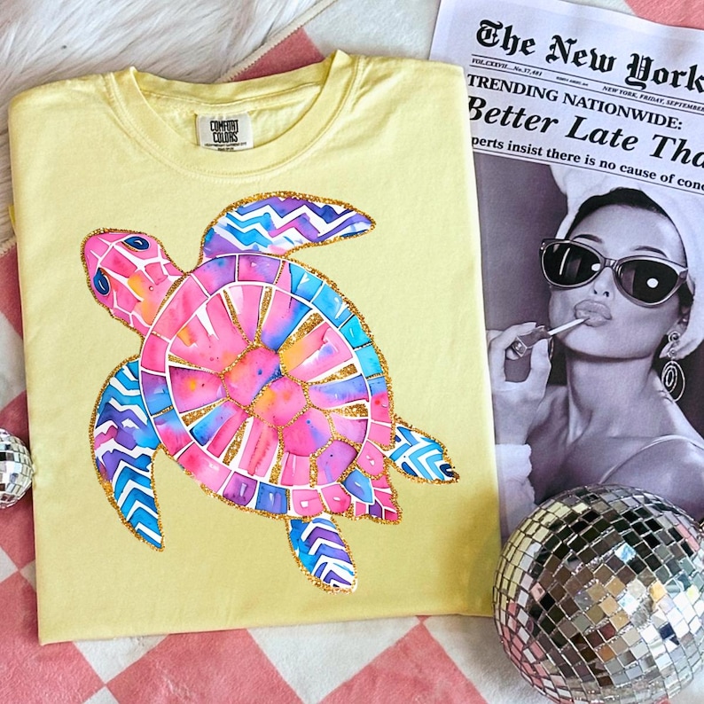 K&ouml;nnte beinhalten: Ein hellgelbes Comfort Colors T-Shirt mit einem farbenfrohen Aquarell-Schildkr&ouml;ten-Design. Die Schildkr&ouml;te hat rosa, blaue und lila Akzente mit goldenen Glitzerdetails. Eine Zeitung und eine Discokugel befinden sich im Hintergrund.