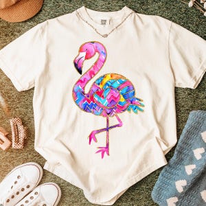 May include: Cream-colored t-shirt featuring a colorful flamingo design. The flamingo has a pink head and neck, a black beak, and a body adorned with a vibrant pattern of blue, yellow, and pink zigzags. The shirt is a comfortable fit.