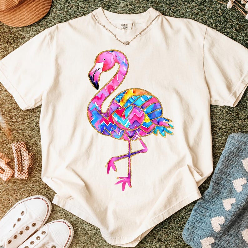 May include: Cream-colored t-shirt featuring a colorful flamingo design. The flamingo has a pink head and neck, a black beak, and a body adorned with a vibrant pattern of blue, yellow, and pink zigzags. The shirt is a comfortable fit.