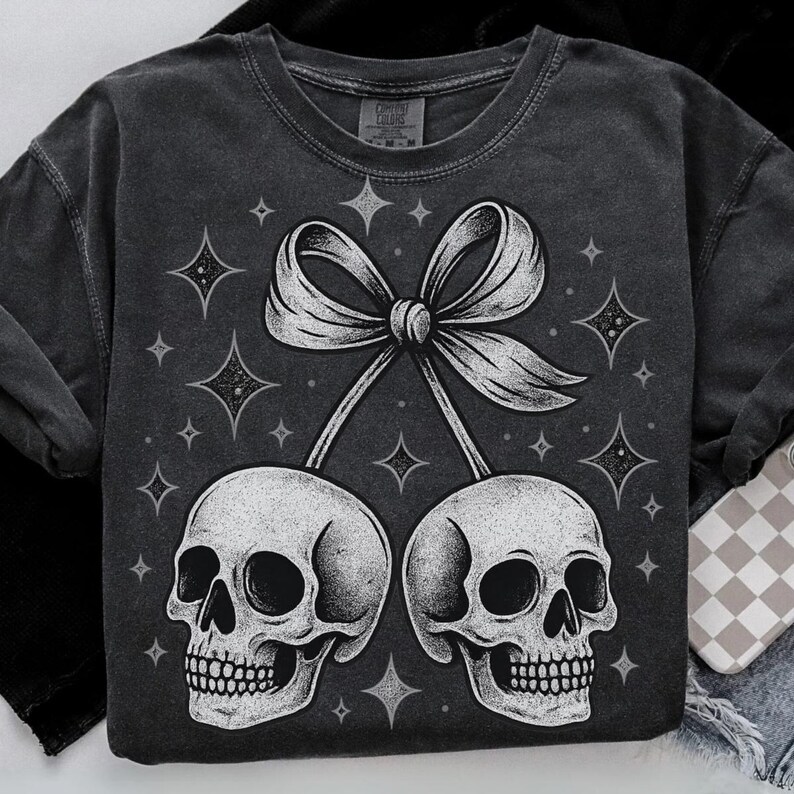 K&ouml;nnte beinhalten: Dunkelgraues Sweatshirt mit einer wei&szlig;en Illustration von zwei Sch&auml;deln, die durch eine Schleife verbunden sind und an Kirschen erinnern. Das Design umfasst funkelnde Sternakzente. Das Sweatshirt ist von der Marke Comfort Colors.