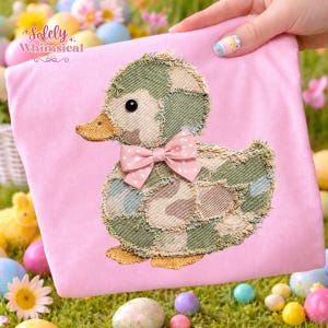 May include: A pink fabric item featuring an embroidered duck design. The duck has a patchwork camouflage body, a pink bow with white polka dots, and a yellow beak. The text "Solely Whimsical" is in the upper left corner.