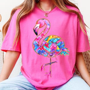 May include: A pink t-shirt featuring a colorful flamingo design. The flamingo has a pink head and neck, a multi-colored body with blue, yellow, and purple patterns, and gold glitter accents. The shirt is a solid pink color.