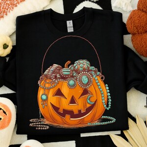 May include: Black sweatshirt featuring a smiling jack-o'-lantern with turquoise and silver jewelry. The pumpkin has a handle and is filled with more jewelry. A crescent moon charm hangs from the pumpkin.