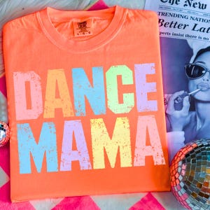 May include: An orange t-shirt with the words "DANCE MAMA" in large, distressed, multi-colored letters. The shirt is made of a soft material and has a crew neck. The shirt is displayed on a pink and white patterned surface.