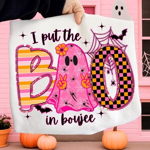 May include: White Halloween-themed blanket with the phrase "I put the BOO in boujee." The design features a pink ghost with peace signs, spiders, and a flower, along with a black and yellow checkered "O".