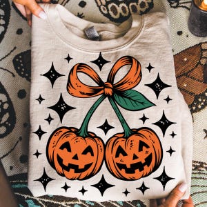 May include: Beige sweatshirt featuring a Halloween-themed design. The graphic includes two orange jack-o'-lanterns connected by green stems and an orange bow, surrounded by black starbursts. The sweatshirt is folded, with the design visible.